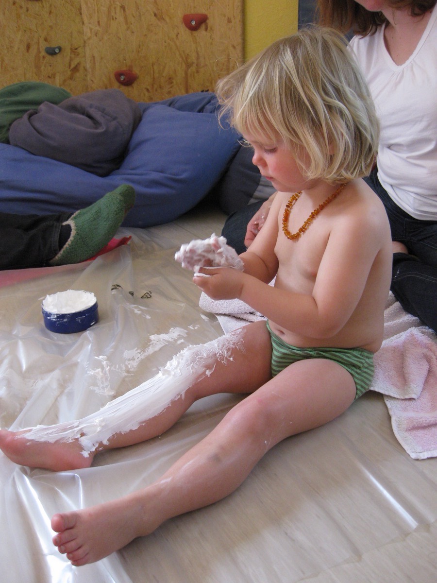 Ein Kleinkind sitzt auf einer Folie und verteilt mit den Händen Creme auf dem eigenen Bein.