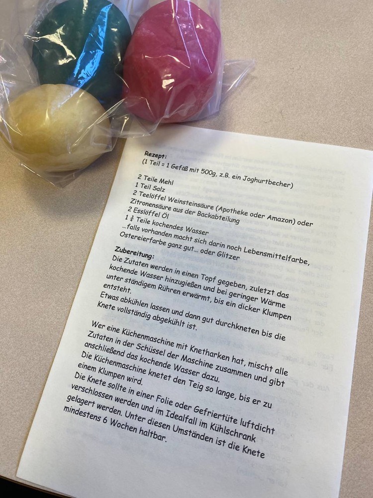 Drei Kugeln selbstgemachte Knete in Gelb, Grün und Pink, eingepackt in Frischhaltefolie, neben dem Rezeptblatt.