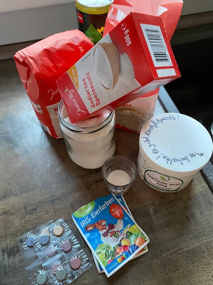 Die Zutaten auf einem Tisch: Mehlpackung, Salz im Schraubglas, Weinsteinsäure, Ostereierfarben und ein Messbecher.